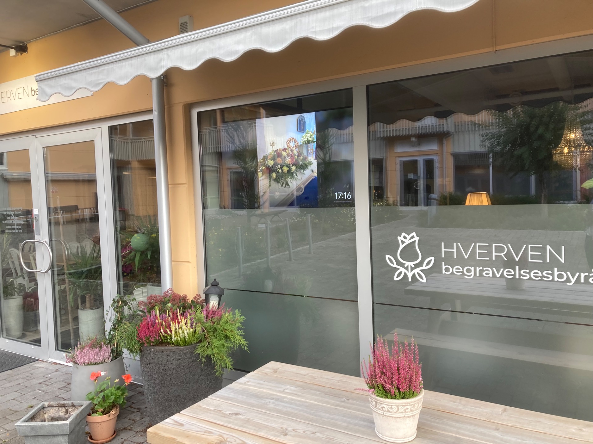 Inngang til HVERVEN begravelsesbyrå med glassfasade, blomsterkasser og hvite markiser over vinduer.